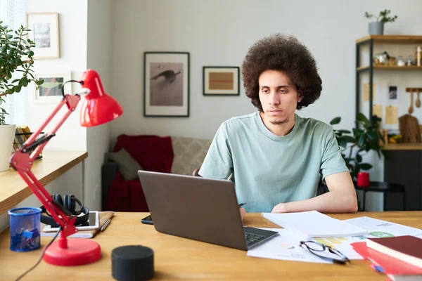 家で文書や他のガジェットとテーブルに座っている間 若い男はノートパソコン上でオンラインで作業 — ストック写真