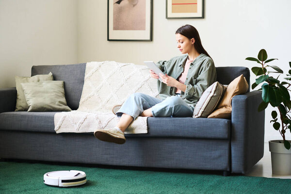 Young woman using digital tablet while robot vacuum cleaner cleaning the carpet in the room