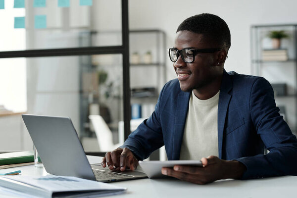 Young smiling businessman in eyeglasses looking at laptop screen while analyzing online data or communicating in video chat in office