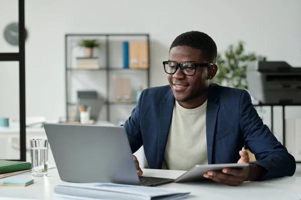 年轻的成功的非裔美国商人 戴着眼镜 穿着雅致的西服 坐在工作场所的笔记本电脑前 用移动小玩意 — 图库照片