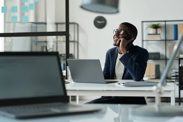 快乐的非洲裔美国经纪人坐在开放空间办公室的笔记本电脑前 一边用智能手机与客户交谈 — 图库照片