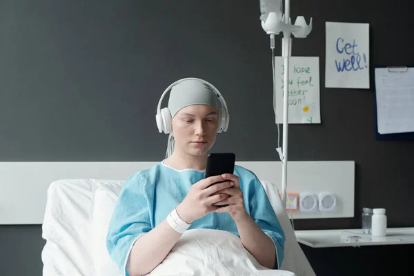 Young serious woman with headphones watching online video or texting in smartphone while sitting in bed in hospital ward after medical treatment
