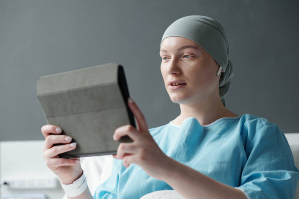 Young bald woman with cancer watching online video in tablet or communicating in video chat with her family or friends in clinics