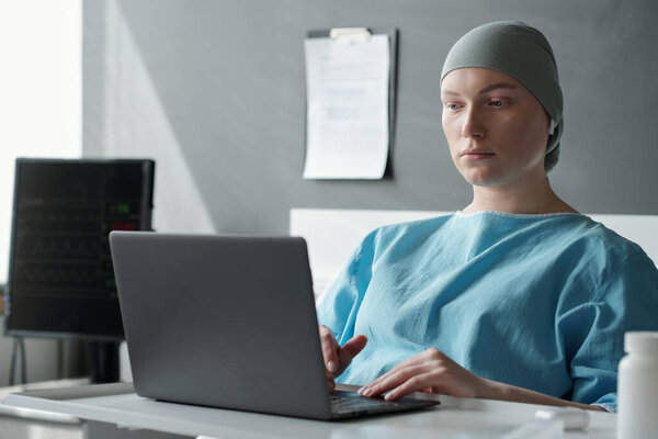 Young serious woman with oncological disease looking at laptop screen while sitting in hospital ward and communicating in video chat