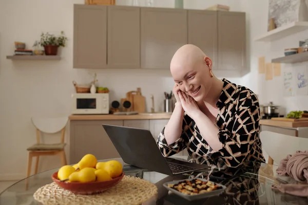 stock image Happy young bald woman looking at laptop screen while sitting by table in the kitchen and watching online movie or communicating with friend