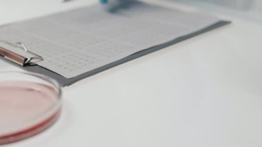 Close-up of foodtech researcher looking through scientific document on desk and putting petri dish with piece of lab-grown meat on his workplace during experiment