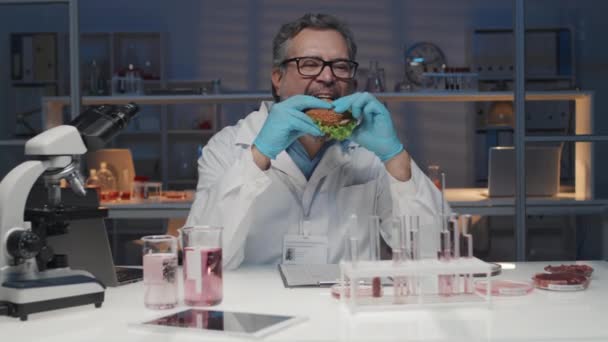 満足した成熟した男性科学者 ワークウェアで おいしいハンバーガーを噛む 実験室で職場で座ってランチを楽しんでいる間 — ストック動画