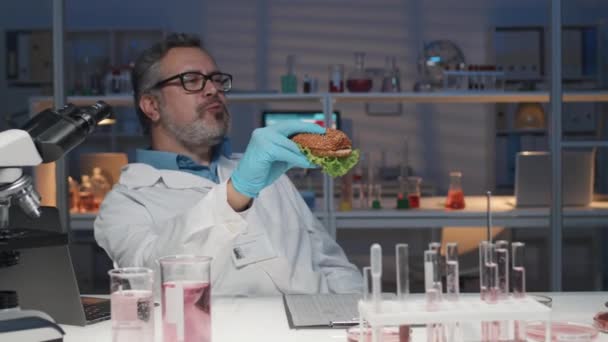 ハッピー成熟した男性研究者は 顕微鏡とテストチューブで机に座って ランチ休憩中にファーストフードを楽しんでいる間 ラボで栽培された肉と美味しいハンバーガーを楽しんでいます — ストック動画