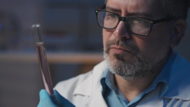 实验室科学实验期间 眼镜中认真的男性基因工程人员凝视着装有液体和微小栽培植物幼苗的瓶中的特写 — 图库视频影像