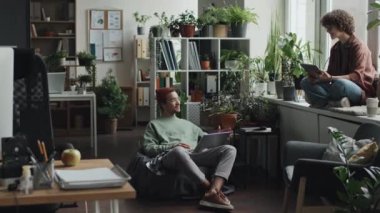 Çevrimiçi projeleri tartışırken yeşil bitkilerin arasında modern ofiste oturan genç meslektaşların geniş açılı görüntüleri.