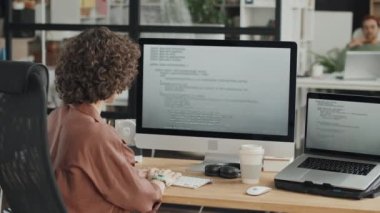 Arkadaki görüntüde genç bir kadın büyük ekranın önünde oturuyor ve kodlarla çalışıyor.
