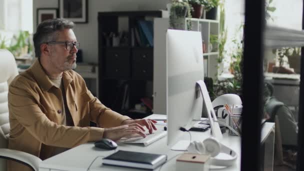 成熟的It专家坐在他的工作场所 看着电脑显示器 在键盘上打字 他与在线项目一起工作 — 图库视频影像
