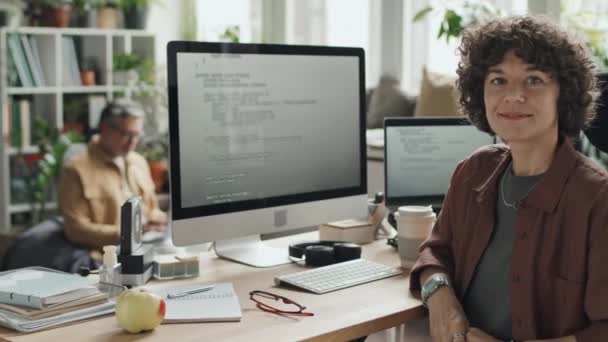 モニターのコード付きコンピュータで彼女の職場に座ってカメラで微笑む若いItスペシャリストの肖像 — ストック動画