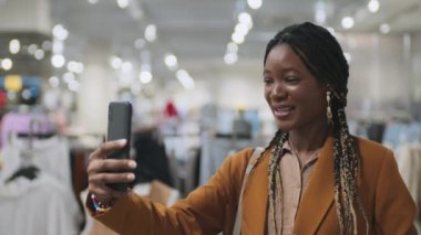 Alışveriş merkezinde alışveriş yaparken akıllı telefonunu kullanarak arkadaşıyla internetten konuşan Afrikalı Amerikalı bir kızın orta boy yakın çekimi.