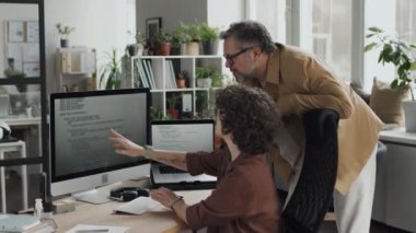 Bilgisayar kodlarını tartışan iki programcı modern ofisteki bilgisayar monitörünü işaret ediyor.