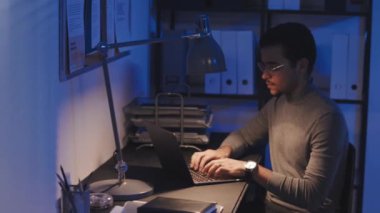 Genç müteahhitin iş yerinde oturup gece geç saatlere kadar dizüstü bilgisayarla çalıştığı orta boy bir fotoğraf.