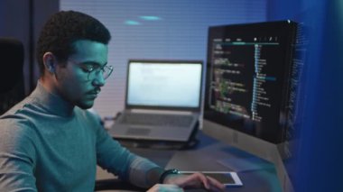 Genç programcı işyerindeki bilgisayar monitörleriyle çevrimiçi işine odaklanıyor, yazılım için kodlar yazıyor.