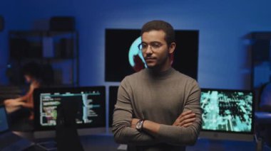 Kolları çapraz duran ve arka planda monitörlerle karanlık bilişim ofisinde dikilirken kameraya bakan genç programcının portresi