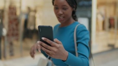 Afro-Amerikalı bir kadın alışveriş merkezinde vitrinlerde yürürken akıllı telefondan konuşuyor.