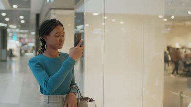 Alışveriş merkezinde alışveriş yaparken akıllı telefonuyla mankenin üstünde fotoğraf çeken Afro-Amerikalı genç bir kadın.