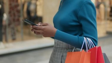 Alışveriş çantalarıyla yakın çekim. Vitrinler boyunca yürürken cep telefonundan mesaj yolluyor.