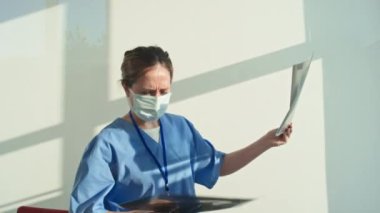 Tanımlanamayan mavi üniformalı ve koruyucu maskeli bir kadın hastanede röntgen görüntülerini okuyor.