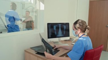 Meslektaşı hastaya danışmanlık yaparken masasında oturan kadın radyolog röntgen görüntüleriyle çalışıyor.