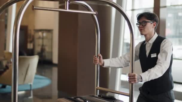 スーツケースを輸送するために荷物トロリーを使用して近代的なホテルポーターやベルホップとして働く若いアジア人男性のスローモ — ストック動画