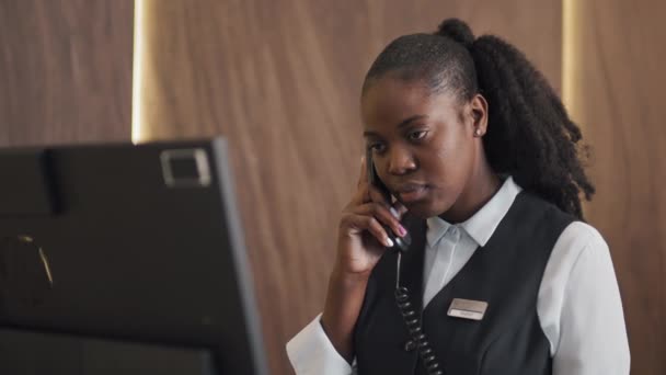 若いアフリカ系アメリカ人の若い女性の肖像画は 新しいゲストと電話で話すホテルのフロントデスクで働く制服を着ています — ストック動画