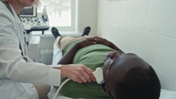 若い黒人の大人の甲状腺の超音波スキャンを行う現代診療所で働く上級白人女性 — ストック動画