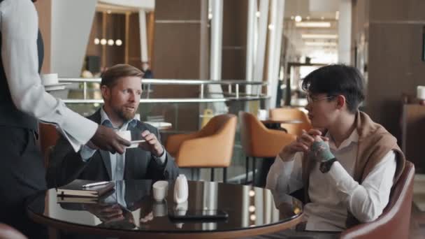 年轻的亚洲男子和他的白人商业伙伴坐在酒店餐厅讨论项目 招待给他们端来咖啡 — 图库视频影像