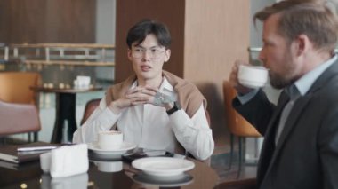 Asyalı genç bir adam şık giyinmiş kafeterya masasında beyaz iş ortağıyla oturup iş hakkında konuşuyor ve kahve içiyor.
