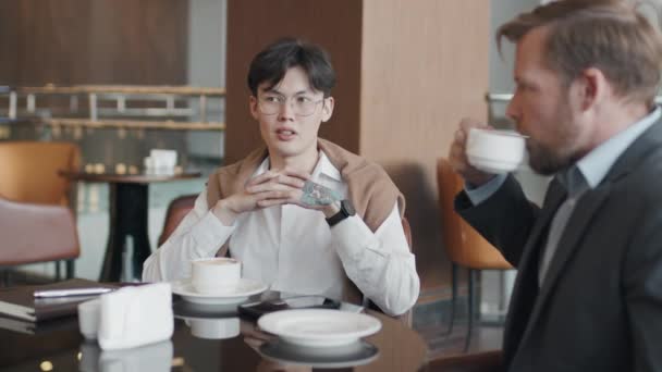 カフェテーブルに座っているスタイリッシュな服を着た若いアジア人男性とコーカサスビジネスパートナーが仕事とコーヒーを飲むことを議論 — ストック動画