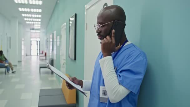 非洲裔美国人医生站在医院走廊上通过电话交谈 他的白人女同事打断了他 问他有关X光拍摄的问题 — 图库视频影像