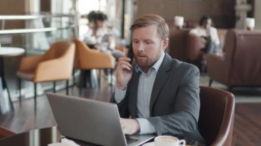 Kafe masasında oturan, yemek molasında laptopla konuşan ve telefonda konuşan resmi giysili beyaz bir genç.
