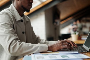 Genç Afrikalı Amerikalı erkek ekonomistin ofis başında dizüstü bilgisayarın önünde otururken internetteki finansal verileri analiz edişinin görüntüsü.