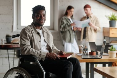 Tekerlekli sandalyede oturan engelli genç, Afro-Amerikan erkek komisyoncu tavan arasında kameranın önünde oturmuş sana bakıyor.