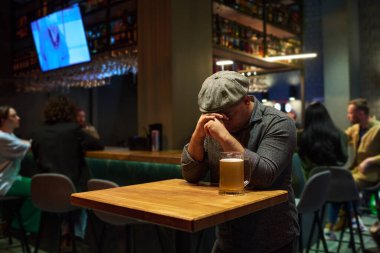 Üzgün, üzgün ya da endişeli hisseden genç bir adam barda elinde bir kupa birayla masanın üzerine eğiliyor.