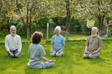 Genç yoga eğitmeni, yeşil çimlerde oturan üç yaşlı insan grubunun önünde sabah eğitimi sırasında huzurevinde yaşıyor.