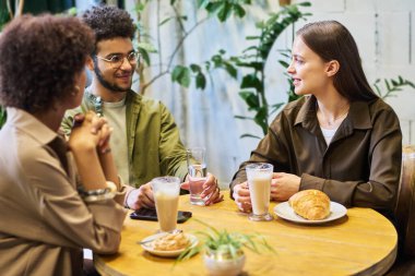 Bir bardak latte ile gülümseyen genç kadın sohbet sırasında sakallı adama bakıyor ve ikisi de rahat bir kafede bayan arkadaşının önünde oturuyor.