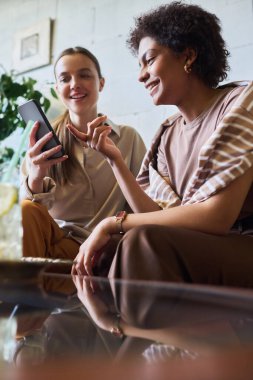 Genç, gülümseyen bir kadın arkadaşının elinde tuttuğu akıllı telefonu işaret ederek modern kafede kameranın önünde oturmuş eğleniyor.