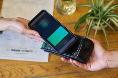 Garsonun elinin üzerinde terminal ve akıllı telefonlu müşterinin kafeteryadaki yiyecek ve içecekler için temassız ödeme sırasında masanın üzerinde durması.