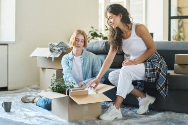 Genç esmer kadın karton kutudan erzakla birlikte saati çıkarırken kanepede oturup kız arkadaşının bavulunu boşaltmasına yardım ediyor.