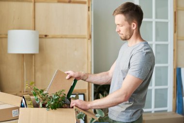 Gri pamuklu tişörtlü genç adam eşyalar paketlenirken ya da paketleri açarken karton kutulara kitap koyuyor.
