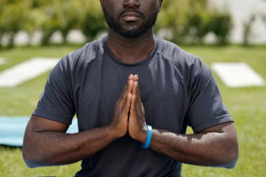 Tişörtlü genç Afro-Amerikan bir adamın yoga egzersizi sırasında kameranın önünde otururken avuçlarını göğsünden tutarken kesilen görüntüsü.