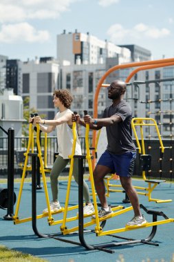 Kültürler arası genç bir kadın ve erkeğin spor tesislerinde antrenman yapması, kollarını ve bacaklarını şehirdeki yüksek binalara dayaması.