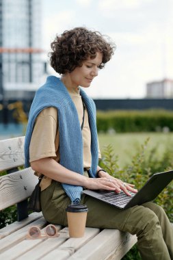 Genç bayan serbest giyimli, parkta ya da şehir ortamında bankta oturuyor ve bilgisayar ekranına bakıp yazarken iletişim kuruyor.