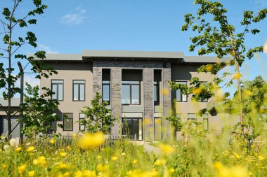 Yeşil çimenlerin, ağaçların ve diğer bitkilerin önünde sıralanmış pencereleri olan gri renkli iki katlı bir evin cephesi.