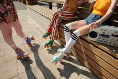 Güneşli bir yaz gününde parkta paten kayarken ahşap bankta oturup sohbet eden üç kadın patencinin yakın çekimi.