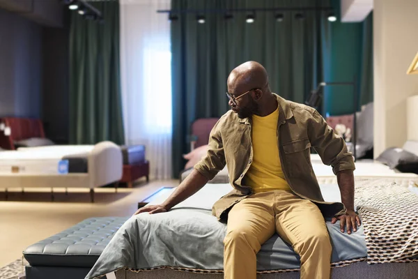 Young black man in casualwear touching blanket while sitting on double bed in furniture shop or department while choosing new item for sleep